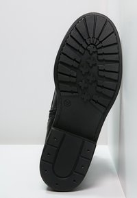 Semelle de chaussure noire avec un motif de crampons profonds, comprenant des lignes horizontales et verticales, une surface texturée et un design à bout arrondi. Taille marquée 37.