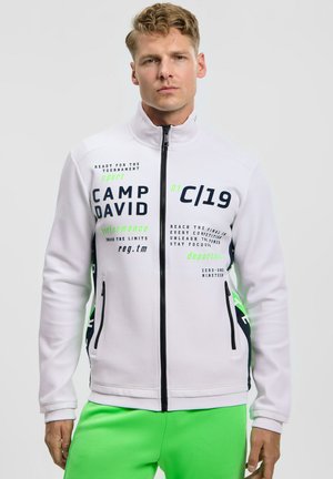 Junger Mann trägt eine weiße Jacke mit Reißverschluss, bedruckt mit schwarzem und grünem Text, sowie leuchtend grüne Hosen, steht vor einem schlichten weißen Hintergrund.