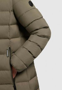 Puffer jacket in olive green, featuring a quilted design, zippered pocket, and a round black logo patch on the shoulder. Smooth texture.