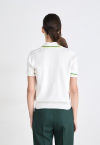 Witte polo met korte mouwen, groene en gele kraagafwerking, ribgebreide onderrand en zijsplitten, gecombineerd met donkergroene op maat gemaakte broek.
