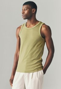 Top de tirantes acanalado en verde oliva con amplias sisas, cuello redondo y diseño ajustado. Combinado con pantalones beige claros, lo que resalta un estilo limpio.