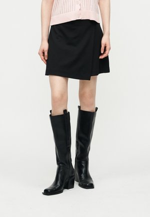 Person iført et svart knelangt skjørt, blek rosa knappet topp og svarte skinnstøvler med hæl som går til knærne, mot en ensfarget bakgrunn.