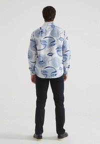 Chemise blanche avec des motifs de visages abstraits bleus et manches longues, associée à un pantalon noir et des chaussures. La vue de dos montre le détail du col.