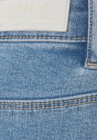 Gros plan sur un jean en denim bleu montrant des coutures doubles et une étiquette de marque en cuir blanc portant l'inscription ONLY.