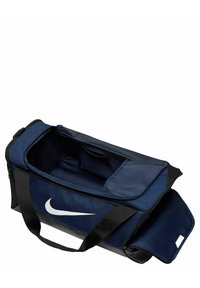 Sac de sport bleu marine avec un logo Nike blanc, doté d'un dessus zippé, de deux poignées et d'une finition en tissu texturé. Inclut une poche latérale.