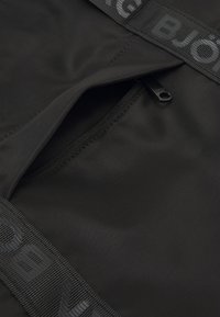 Saco de tecido preto com um bolso com fecho éclair e uma pega de hardware preto lisa. As bordas são acabadas com uma alça de webbing texturizada preta.