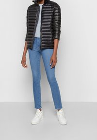 Sort dunjakke med vandret quiltet design, grå top under, kombineret med lyseblå tætsiddende jeans og hvide høje sneakers.