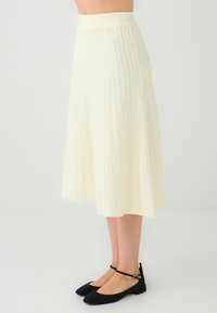 Jupe midi plissée crème à taille haute, réalisée en tissu doux, avec des rayures verticales et une silhouette fluide. Ballerines noires présentées.