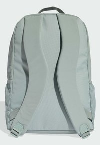 Mochila gris claro con correas acolchadas y bolsillos laterales de malla. Presenta un asa en la parte superior y una tela suave con acentos texturizados.