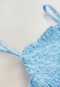 Kék ráncolt felső, amely összegyűrt textúrával és vékony pántokkal rendelkezik, a virágmintát a szövet kialakításába integrálva.