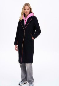 Žena oblečená v čiernom kabáte s farebným lemovaním, pod ktorým má na sebe ružovú mikinu, šedé džínsy a strieborné tenisky, stojí pred bielym pozadím.