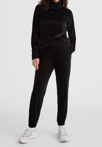 Chándal de terciopelo negro con cuello de capucha y cintura elástica. Mangas largas, bolsillos laterales, combinado con zapatillas deportivas blancas y grises.