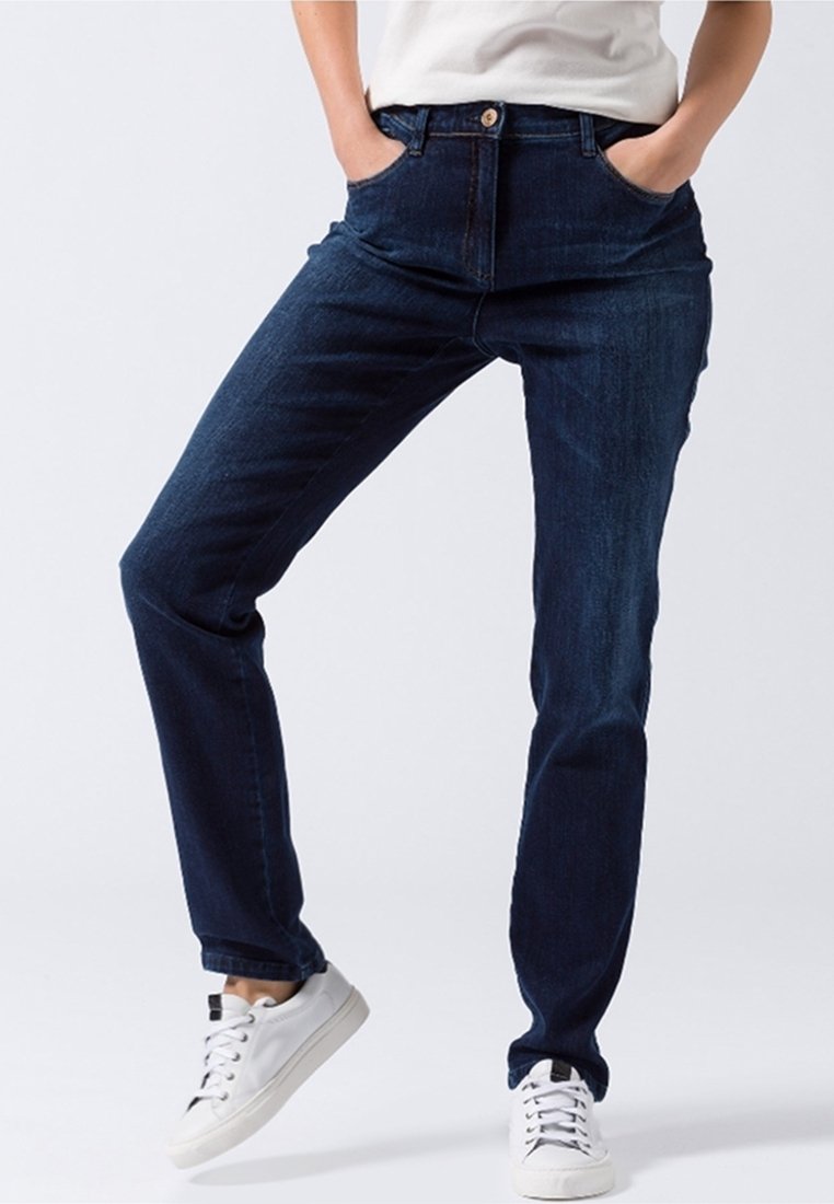 Personne portant un jean bleu foncé et des baskets blanches, debout les mains dans les poches sur un fond clair uni.