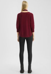 Blonde vrouw staat met haar rug naar de camera gekeerd, gekleed in een bordeauxrode trui, zwarte strakke broek, zwarte laarzen en een donkerbordeauxrode sjaal.