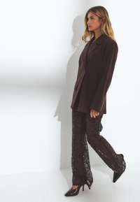 Bruine oversized blazer gecombineerd met doorschijnende zwarte kantbroeken en puntige zwarte hakken. Soepele texturen en op maat gemaakte ontwerpen zichtbaar.