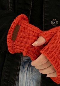 Guanti senza dita rossi lavorati a maglia con texture a coste e una piccola etichetta marrone. Indossati sopra una giacca nera con del tessuto denim visibile sotto.