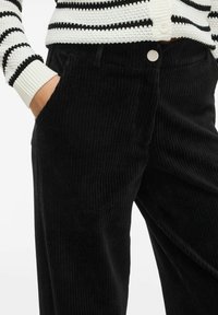 Melna korduroja džinsi ar paceltām vertikālām svītrām, ar sānu kabatu un sudraba pogu aizdari. Virsū ir svītraina adīta džempera.