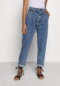 Korkeavyötäröiset, siniset farkuhousut, joissa on vyövyö, kapenevat lahkeet, käärityt hihnat ja hieman haalistunut pinta. Yhdistetty mustiin nauhakengiin.