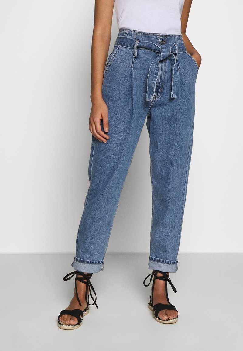 Korkeavyötäröiset, siniset farkuhousut, joissa on vyövyö, kapenevat lahkeet, käärityt hihnat ja hieman haalistunut pinta. Yhdistetty mustiin nauhakengiin.