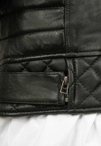 Primo piano di una sezione di giacca in pelle nera trapuntata con cerniera argentata e dettaglio di cinturino in pelle su sfondo di tessuto bianco.