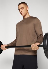 Langermet treningsskjorte i brunt, laget av glatt stoff. Modellen løfter en svart manual iført mørke shorts. Minimalistisk logo på brystet.