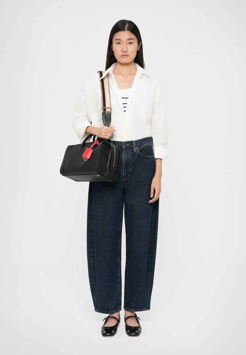 Sort håndtaske med rød detalje og aftagelig skulderrem, båret med en hvid skjorte over en stribet top, parret med vide denim jeans.