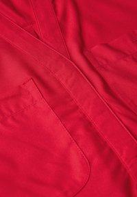 Czerwona tkanina o gładkiej fakturze, z dwoma przednimi kieszeniami i stębnowanym kołnierzykiem. Tkanina ma delikatny połysk i estetyczne wykończenie.