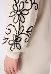 Langarmig kledingstuk in beige met zwarte bloemenborduring op de mouw. Soepele stof en geribbeld manchetdetail. Elegante stijl.