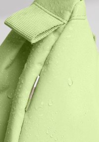 Tessuto verde chiaro con una superficie testurizzata, caratterizzato da gocce d'acqua. Una cucitura visibile e un pezzo di accento di colore chiaro evidenziano il design.