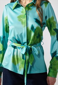 Bluză cu mâneci lungi, de culoare bleumarin deschis, cu un model tie-dye în nuanțe de verde și teal închis. Dispune de un închidere cu nasturi în față și un brâu asortat legat la talie.