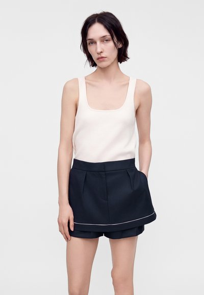 Femme aux cheveux courts et foncés, portant un haut blanc sans manches et un short noir superposé avec une bordure blanche, debout devant un fond uni.