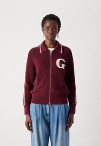 Cardigan a maglia bordeaux con zip frontale, rifinitura bianca del colletto, grande logo "G" bianco sul petto e tasche laterali, abbinato a pantaloni azzurri chiari.