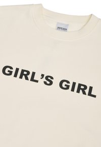 Sweat-shirt à col rond de couleur crème avec un texte noir en gras "GIRL'S GIRL" sur la poitrine et une étiquette de marque visible à l'intérieur du col.