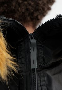 Sort zip-jakke med hætte foret med pels, quiltet indvendig og en sort lynlås med branding synlig ved halsen.