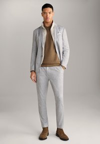 Completo in lana grigio chiaro con giacca strutturata, un maglione testurizzato beige e stivaletti in suede marroni, con uno sfondo neutro.
