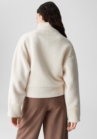 Cremefarbene Fleecejacke mit hohem Kragen, gerippten Bündchen und cropped Design. Weiches, strukturiertes Material. Getragen über braunen Hosen.