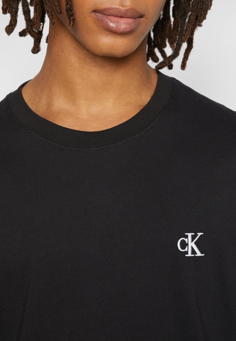 Persona con rastas cortas que lleva una camiseta negra de cuello redondo con un pequeño logo blanco "cK" en la parte superior izquierda del pecho.