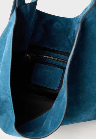 Sac cabas en daim bleu ouvert pour montrer la poche intérieure avec une fermeture éclair visible et une étiquette cousue à l'intérieur du sac.