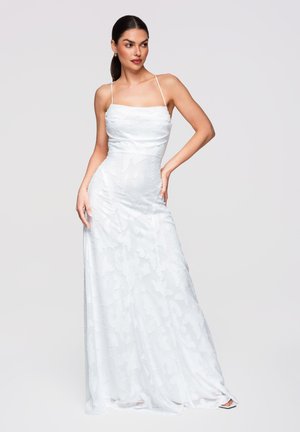 Kobieta pozująca w białej, sięgającej do podłogi, sukience bez rękawów z cienkimi ramiączkami i subtelnymi teksturowanymi kwiatowymi wzorami.