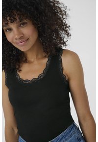 Lächelnde junge Frau mit lockigem Haar, die ein schwarzes Top mit Spitzenrand und blaue Jeans trägt, posiert vor einem neutralen Hintergrund.