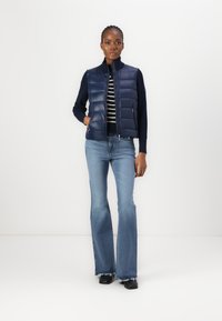 Gilet in nylon blu sopra una maglietta a righe, abbinato a jeans chiari a zampa d'elefante. Texture liscia, design aderente, con tasche con zip.