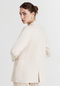 Young woman with dark hair in a low bun wearing cream-colored blazer and matching pants, looking over her shoulder against a plain background.