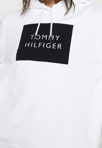 Felpa bianca con cappuccio caratterizzata da una grande stampa rettangolare nera con la scritta "TOMMY HILFIGER" in lettere bianche. Tessuto morbido con cappuccio regolabile.
