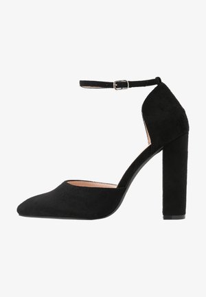 Zapato de tacón alto de ante negro con punta afilada, tira en el tobillo y tacón grueso. Textura suave y diseño minimalista.