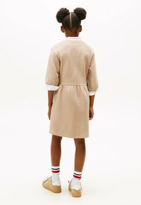 Robe beige côtelée avec une taille cintrée, des manches arrivant aux coudes et un col blanc. Portée avec des chaussures beiges et des chaussettes blanches à rayures rouges et bleues.