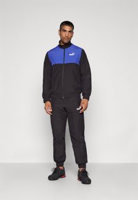 Schwarzer sportlicher Trainingsanzug mit blauem Oberteil, elastischen Bündchen und elastischem Bund. Logo auf der Brust und dem Hosenbein. Schwarze Sportschuhe mit roten Akzenten.