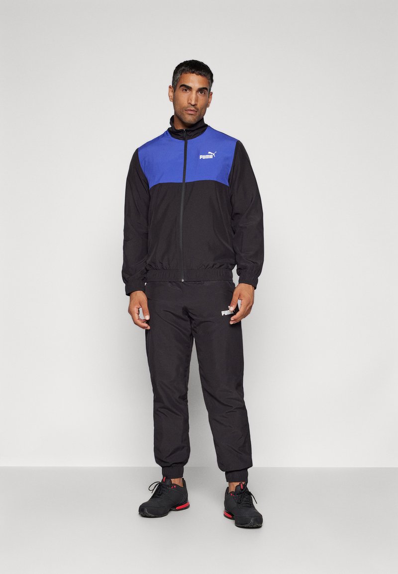 Schwarzer sportlicher Trainingsanzug mit blauem Oberteil, elastischen Bündchen und elastischem Bund. Logo auf der Brust und dem Hosenbein. Schwarze Sportschuhe mit roten Akzenten.