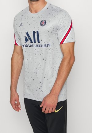 Mann trägt graues Paris Saint-Germain Fußballtrikot mit schwarzen Sprenkeln und roten, weißen Streifen an den Ärmeln.