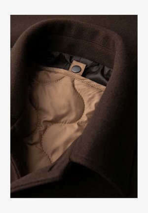 Hnědý vlněný kabát s prošívanou světle hnědou podšívkou. Má vysoký límec a detail se cvočkem u krku. Jemná textura, střih na míru.