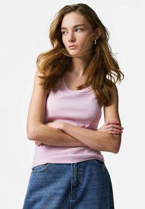 Noor naine pikkade laineliste juustega, seljas roosa varrukateta top ja sinised teksad, seisab käed rüpes ja vaatab kõrvale.